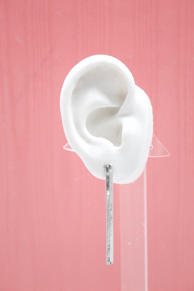 A white mannequin ear displays a sleek, silver herringbone chain drop earring against a textured pink background.