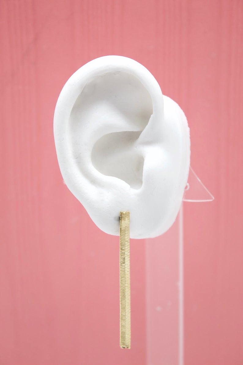 A white mannequin ear displays a minimalist gold bar dangle earring against a solid pink background.