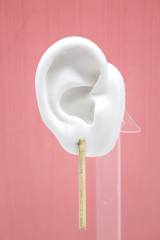 A white mannequin ear displays a minimalist gold bar dangle earring against a solid pink background.