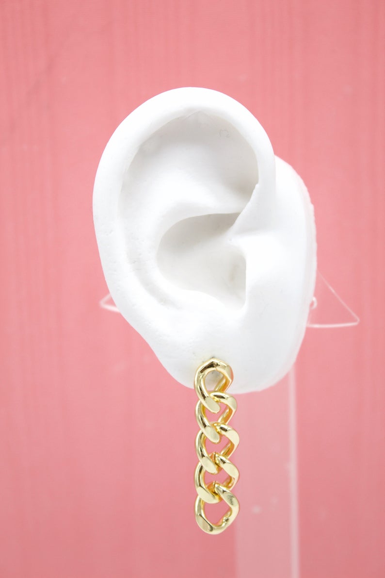 A white mannequin ear displays a dangling gold chain link earring against a pink background.