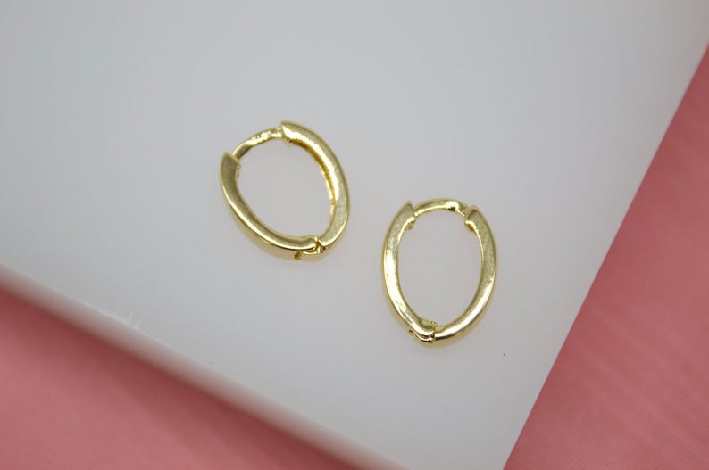 A pair of sleek gold oval huggie earrings rest on a white display surface against a pink background.
