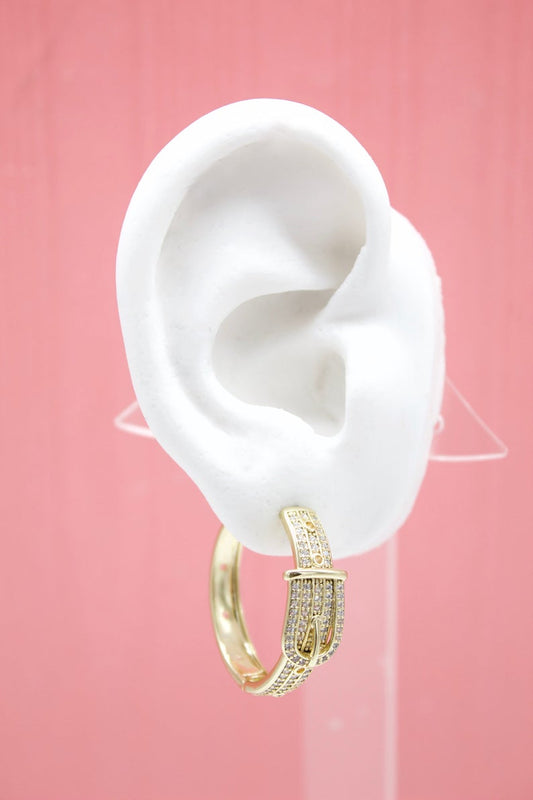 A white ear model displays a gold buckle-shaped hoop earring encrusted with sparkling cubic zirconia stones against a pink background.