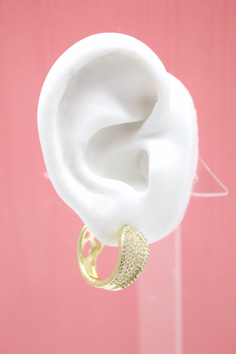 A white mannequin ear displays a sparkling gold huggie hoop earring encrusted with tiny cubic zirconia stones against a solid pink background.