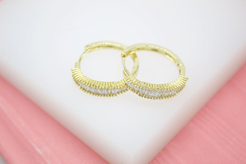 A pair of gold huggie hoop earrings embedded with small white baguette crystals rests on a white and pink surface.