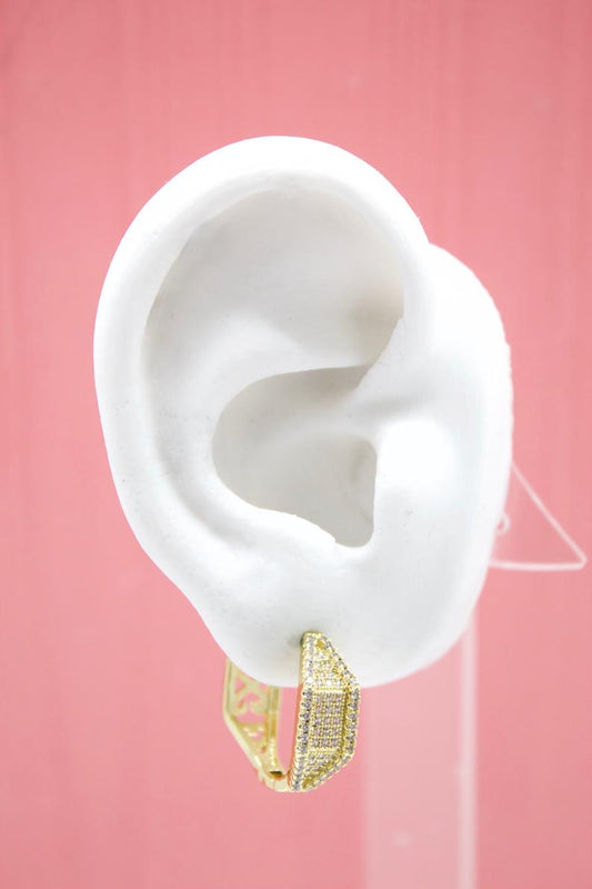 A white display ear model showcases a sparkling gold pave huggie hoop earring against a soft pink background.