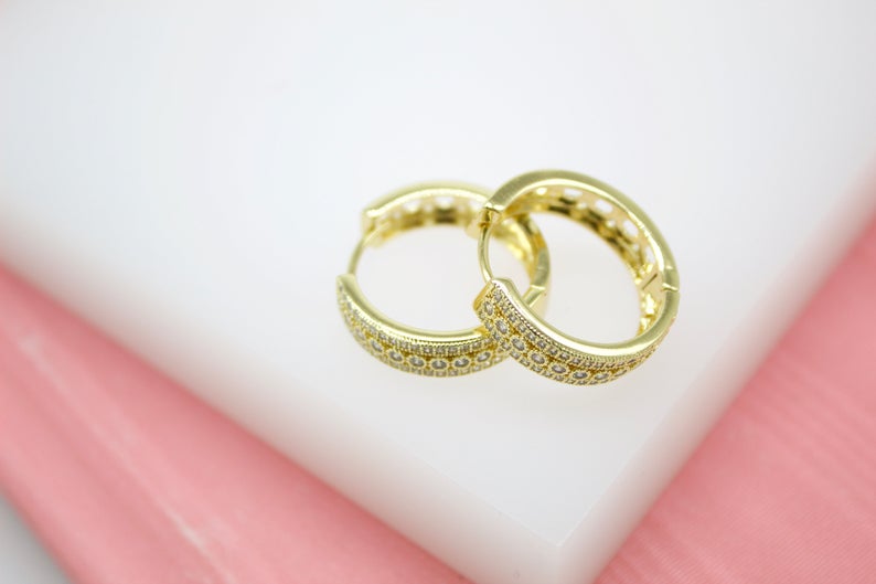 A pair of sparkling gold huggie hoop earrings with pave diamonds rests on a white and pink display.