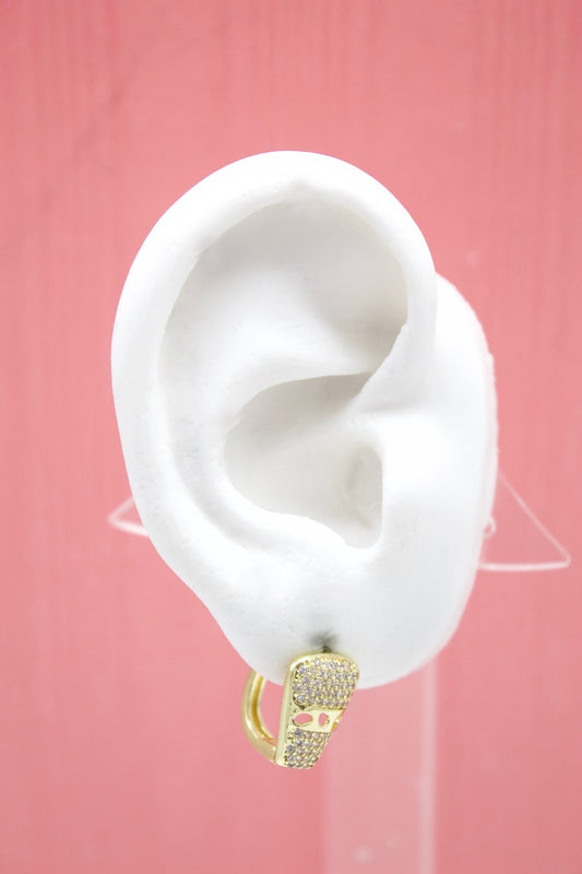 A white mannequin ear displays a sparkling gold skull huggie earring against a pink background.