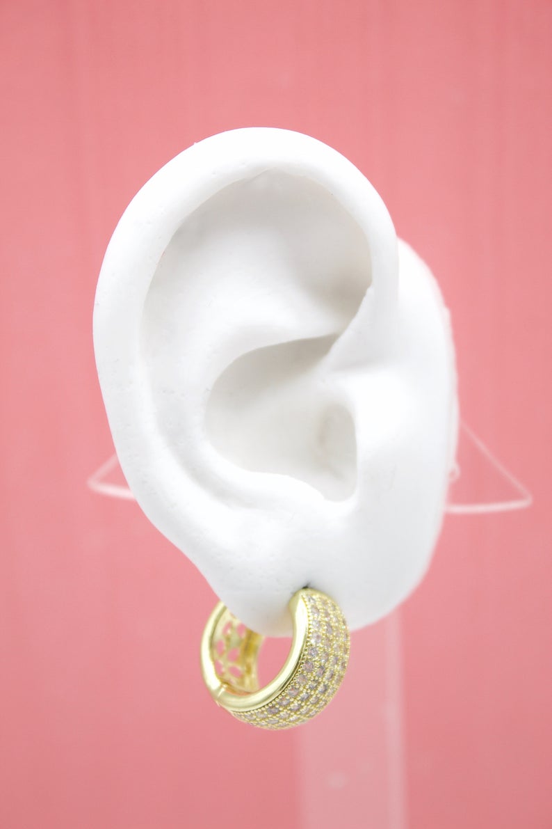 A white mannequin ear displays a thick gold hoop earring encrusted with shimmering cubic zirconia stones against a pink background.