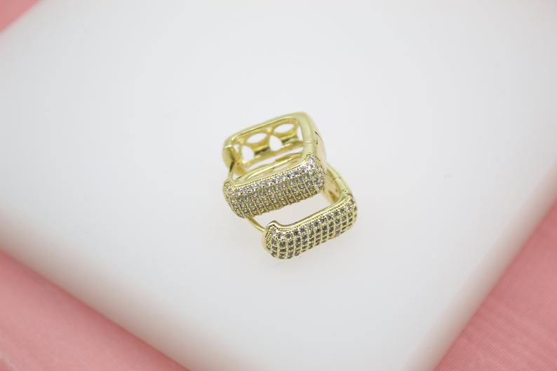 A pair of gold square huggie hoop earrings encrusted with sparkling cubic zirconia pavé rest on a white display surface.