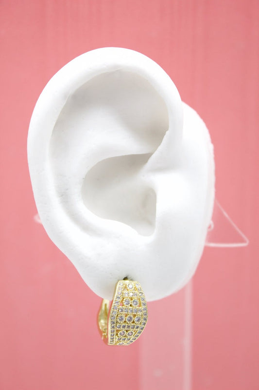 A white mannequin ear displays a wide gold huggie hoop earring encrusted with sparkling cubic zirconia stones against a pink background.