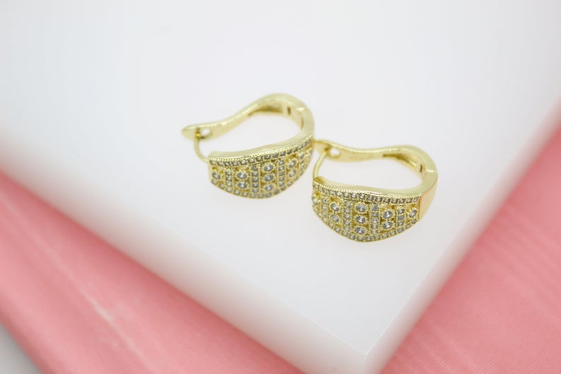 A pair of gold huggie hoop earrings encrusted with sparkling cubic zirconia stones rests on a white display surface.