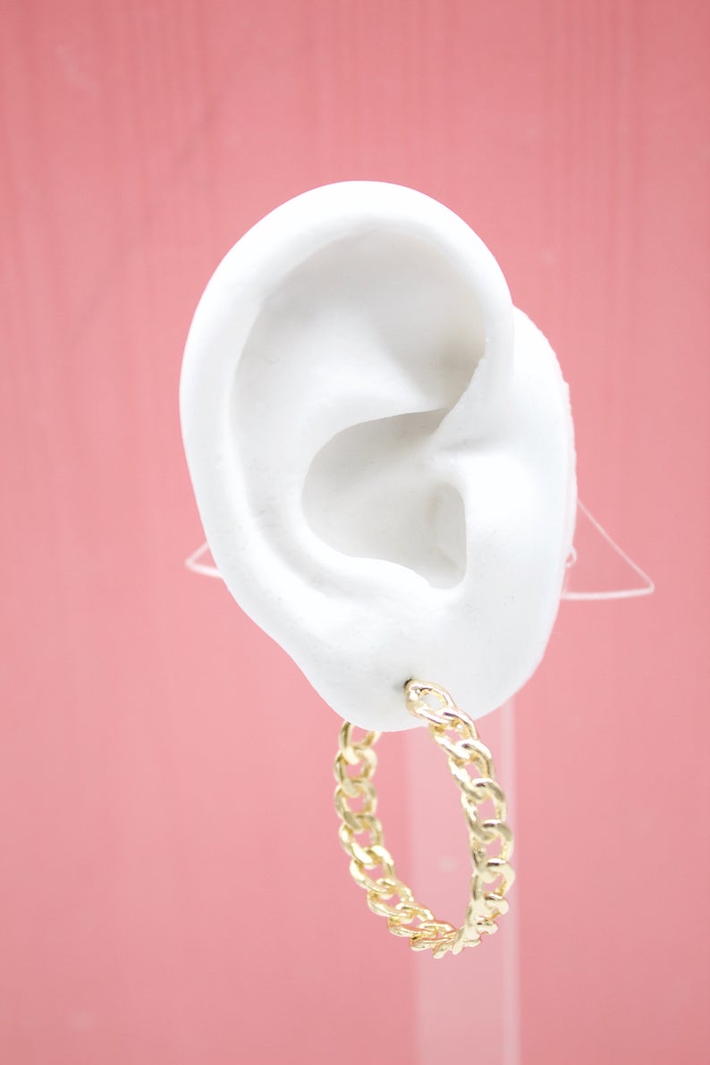 A white mannequin ear displays a stylish gold chain link hoop earring against a pink background.