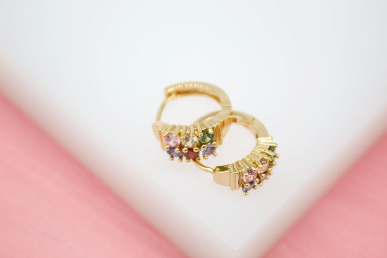 A pair of gold huggie hoop earrings featuring sparkling multicolored cubic zirconia stones rests on a white and pink surface.