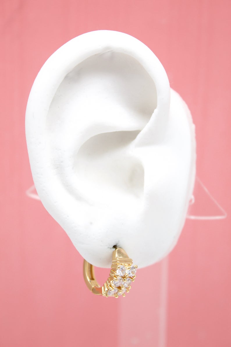 A white mannequin ear displays a gold huggie hoop earring embellished with sparkling cubic zirconia stones against a pink background.