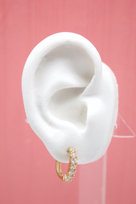 A white ear model displays a sparkling gold hoop earring encrusted with clear gemstones against a soft pink background.
