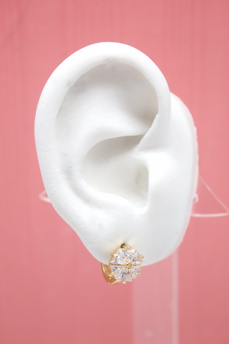 A white mannequin ear displays a sparkling gold stud earring with a central faceted gemstone against a pink background.