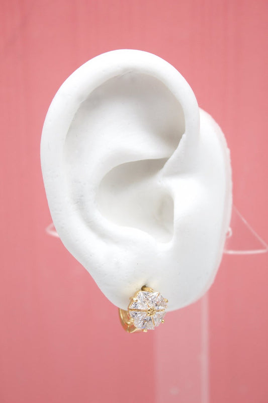 A white mannequin ear displays a sparkling gold stud earring with a central faceted gemstone against a pink background.