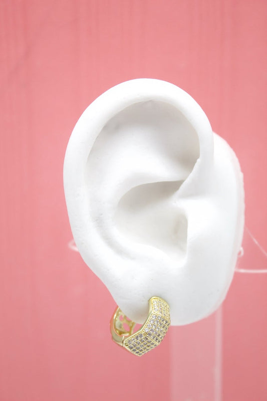 A white mannequin ear displays a gold huggie hoop earring encrusted with small clear crystals against a pink background.