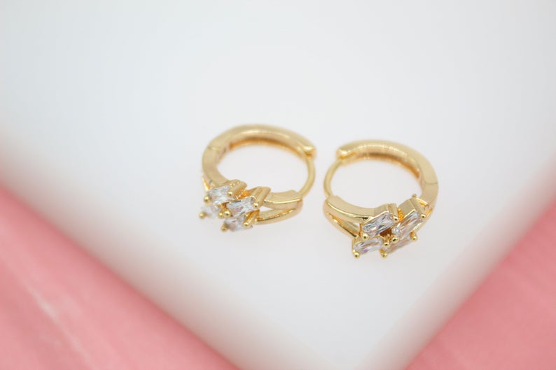 A pair of shiny gold hoop earrings featuring sparkling crystal stones rests on a white and pink surface.