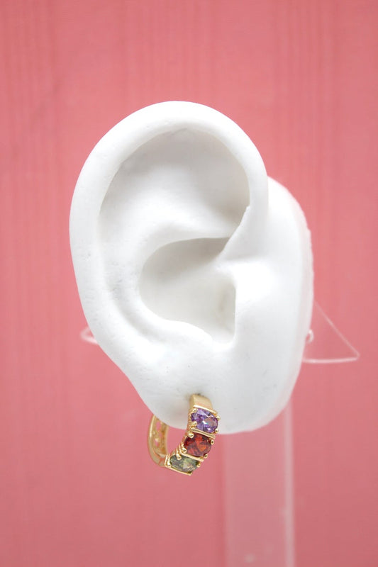 A white mannequin ear displays a gold hoop earring set with vibrant purple, red, and green gemstones against a pink background.