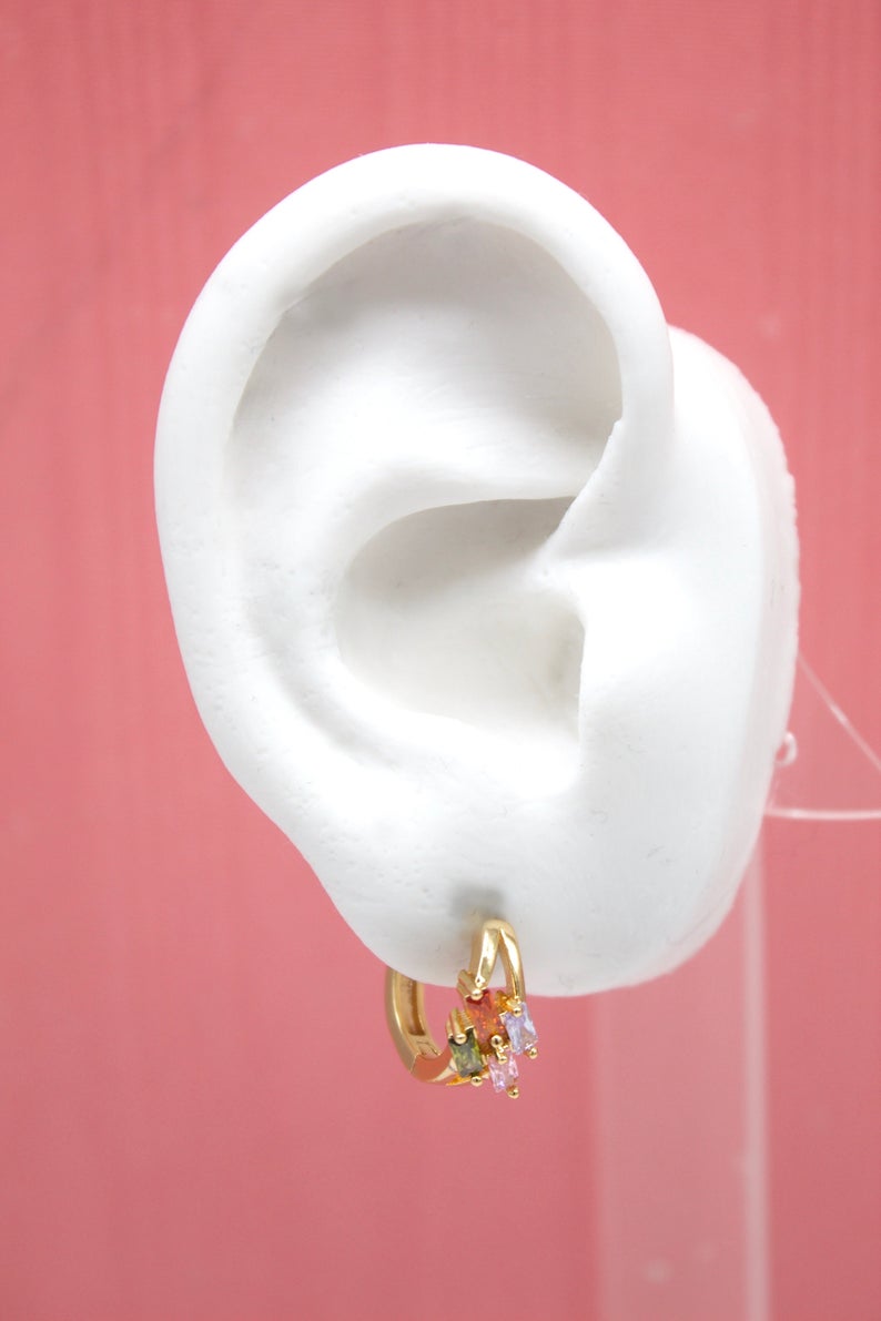 A white mannequin ear displays a gold hoop earring adorned with colorful baguette-cut gemstones against a pink background.
