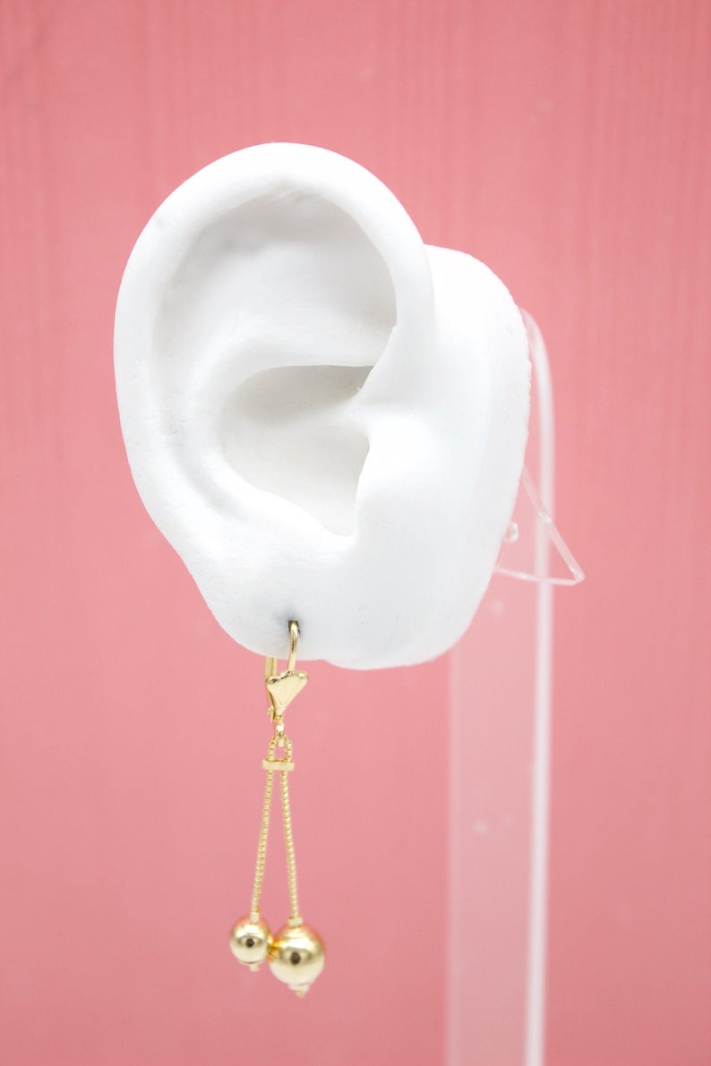 A white mannequin ear displays a delicate gold dangle earring featuring two hanging gold bead charms.