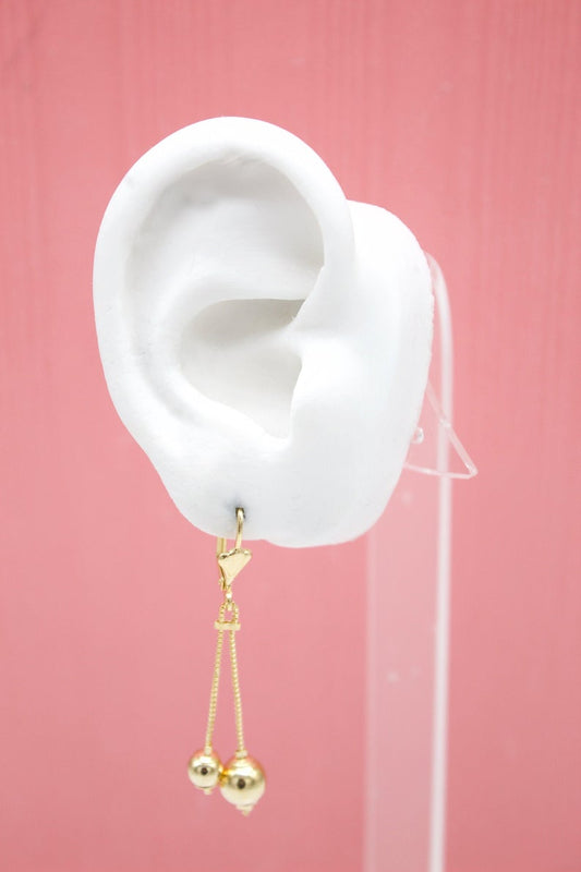 A white mannequin ear displays a delicate gold dangle earring featuring two hanging gold bead charms.
