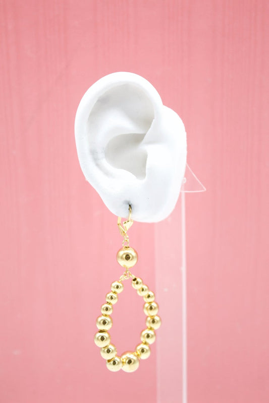 A white mannequin ear displays a dangling gold beaded teardrop earring against a pink background.