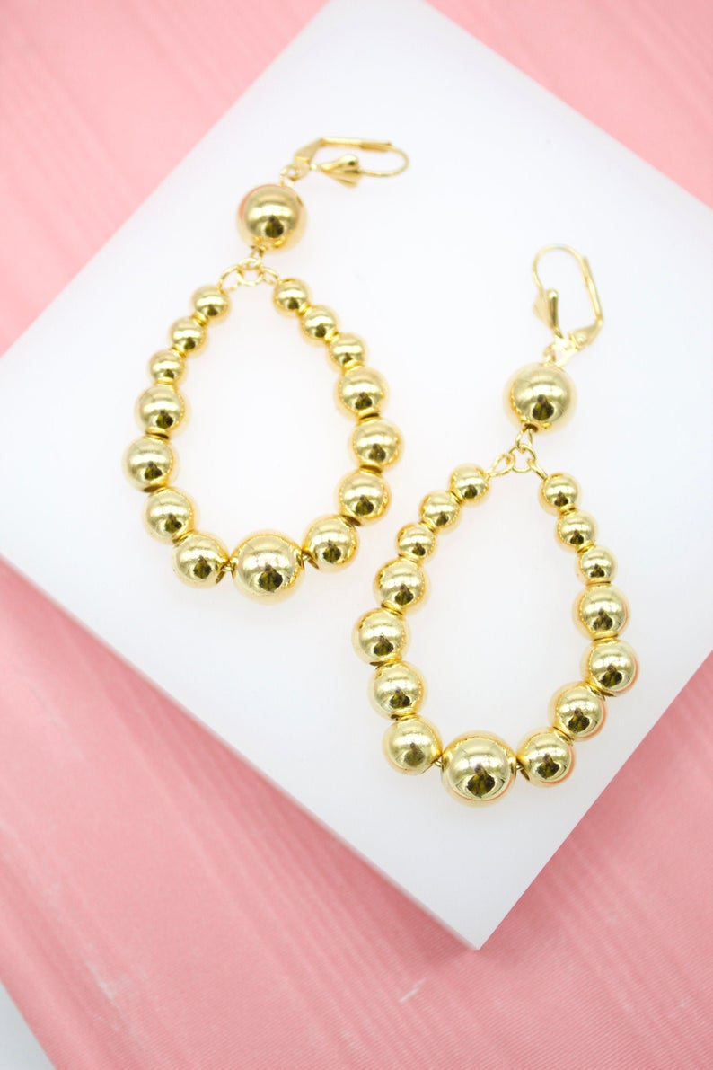 A pair of shiny gold bead drop earrings rests on a white display against a pink background.