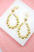 A pair of shiny gold bead drop earrings rests on a white display against a pink background.