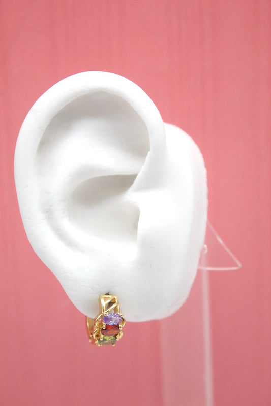 A white mannequin ear displays a gold huggie hoop earring featuring colorful gemstones against a pink background.