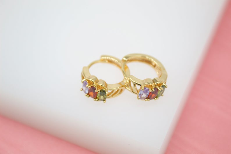 A pair of gold huggie hoop earrings featuring colorful cubic zirconia stones rests on a white and pink surface.