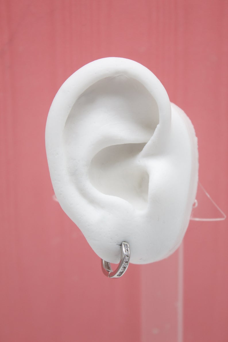A white mannequin ear displays a sparkling diamond huggie hoop earring against a pink background.