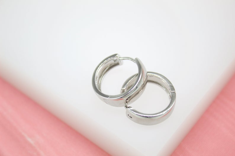 A pair of polished silver huggie hoop earrings rests against a white and pink backdrop.