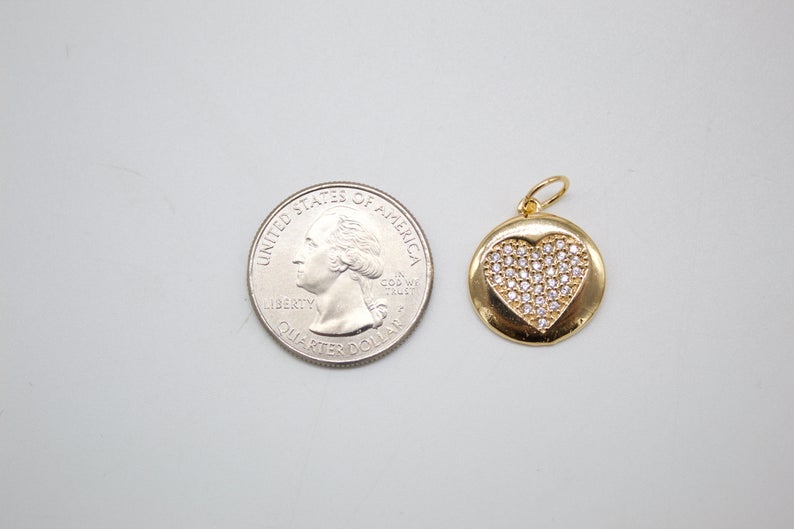 A gold pendant featuring a diamond-pave heart inside a circular frame rests next to a US quarter coin for scale.