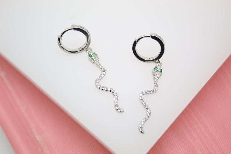 A pair of silver snake hoop earrings features sparkling cubic zirconia bodies and striking green crystal eyes against a white and pink background.