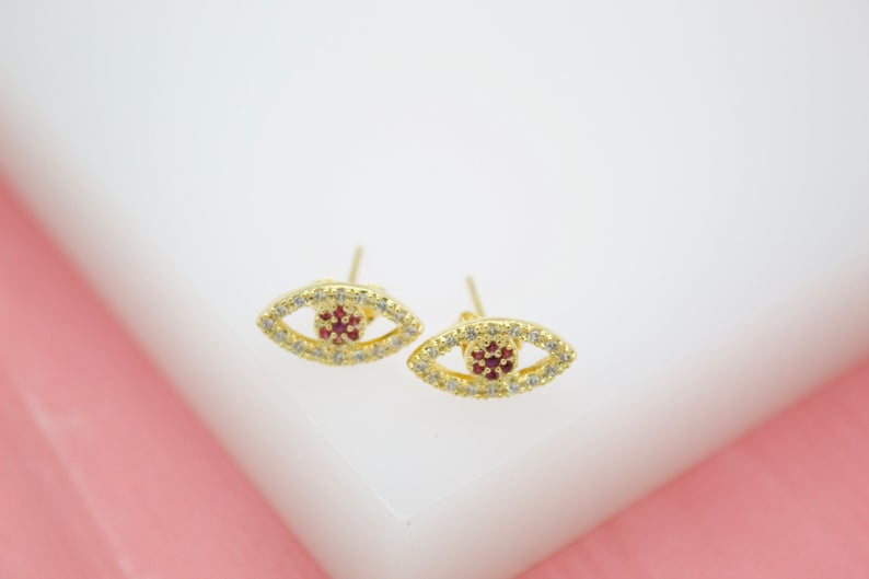 A pair of gold evil eye stud earrings features sparkling pave diamonds and vibrant red gemstone centers against a neutral white backdrop.