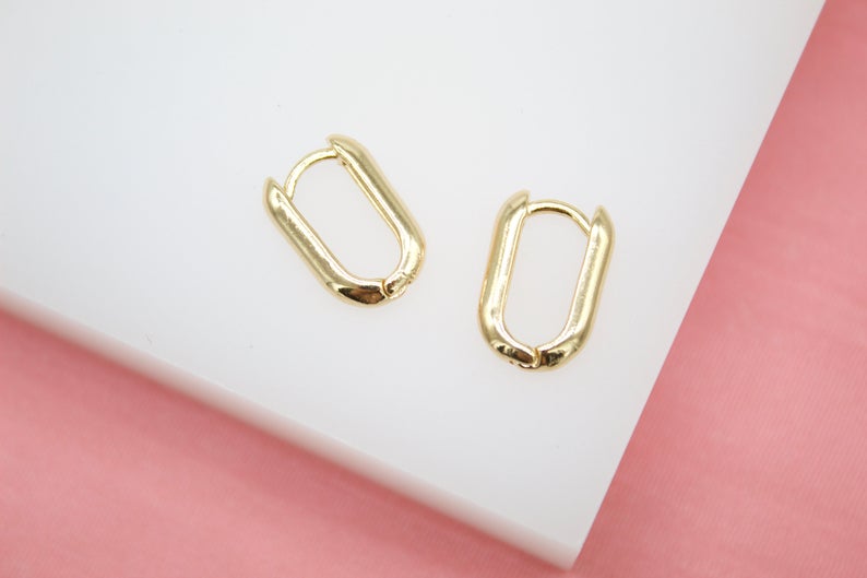 A pair of sleek gold rectangular huggie hoop earrings rests on a white surface against a pink background.