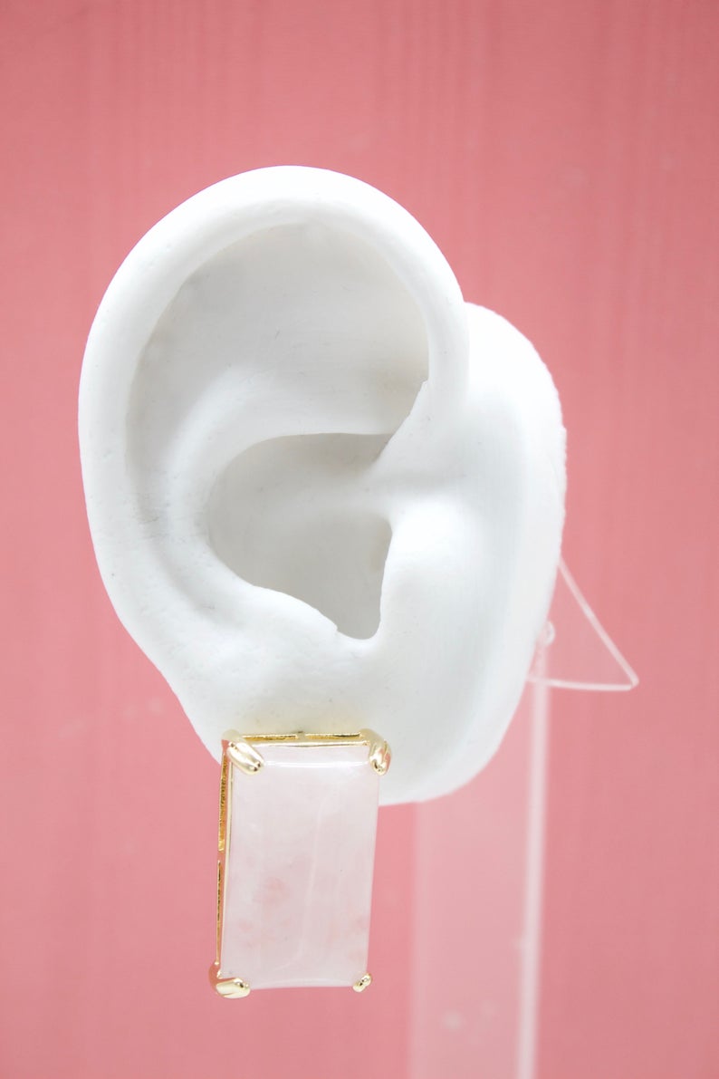 A white model ear showcases a rectangular rose quartz gemstone earring set in gold.