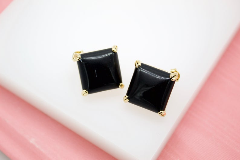 A pair of square black onyx stud earrings rests on a white display surface against a pink background.
