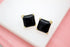 A pair of square black onyx stud earrings rests on a white display surface against a pink background.
