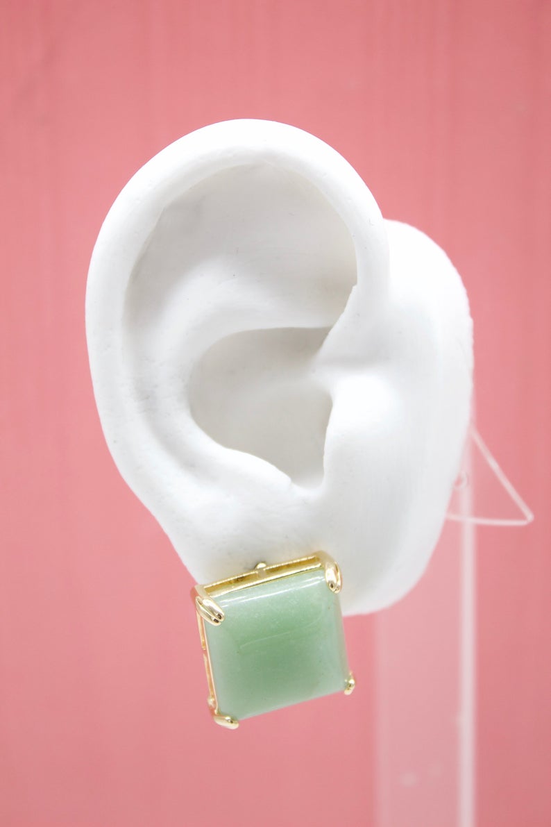 A white mannequin ear displays a square green jade gemstone earring set in gold.