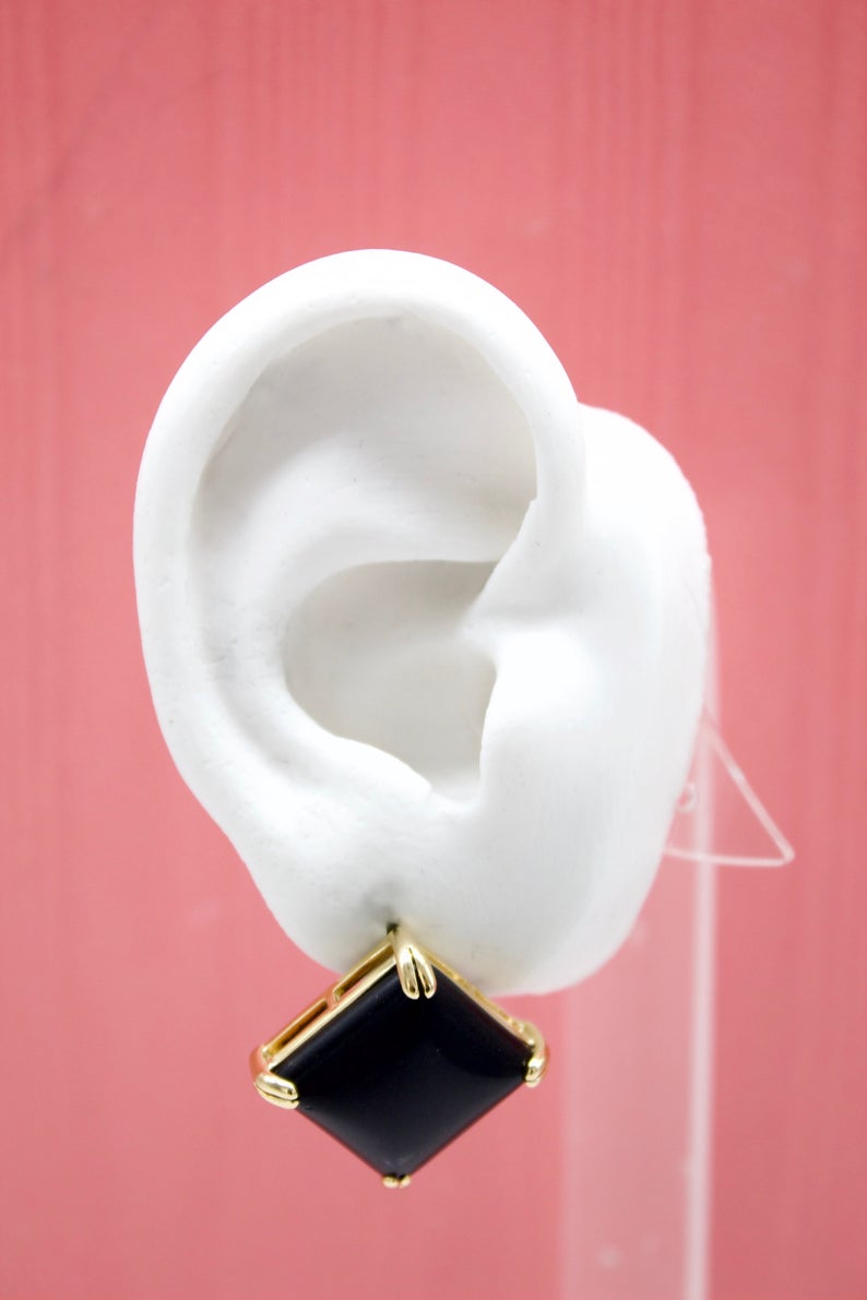 A white mannequin ear displays a square black gemstone earring with a gold setting against a pink background.
