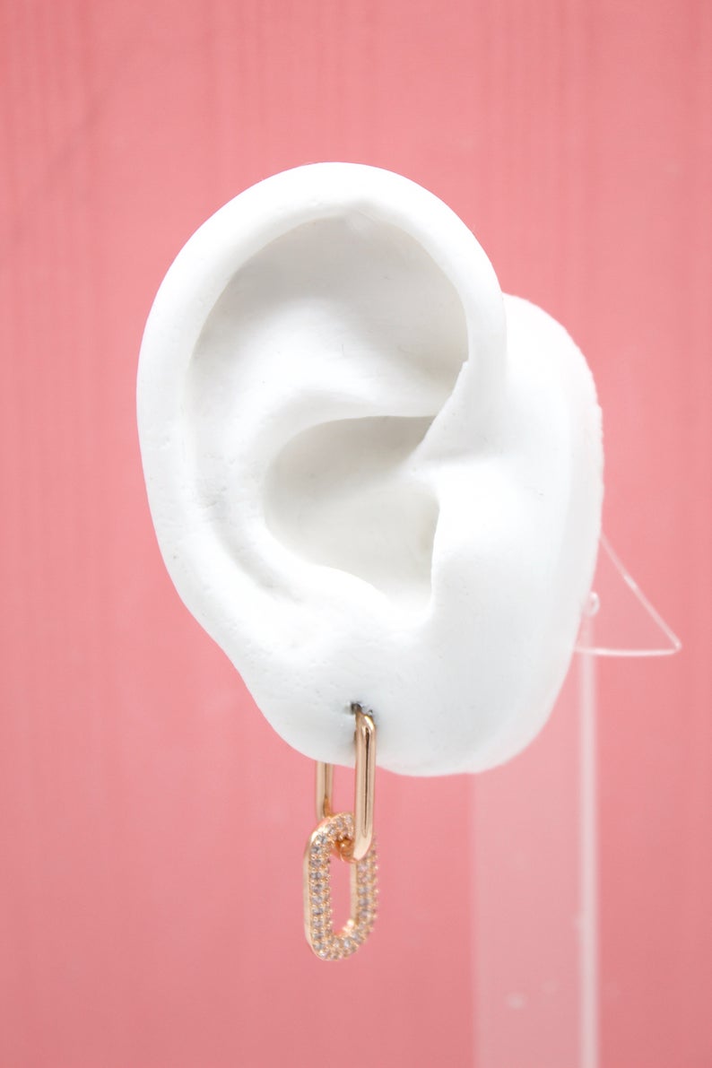 A white mannequin ear displays a gold link earring with sparkling pave diamonds against a soft pink background.