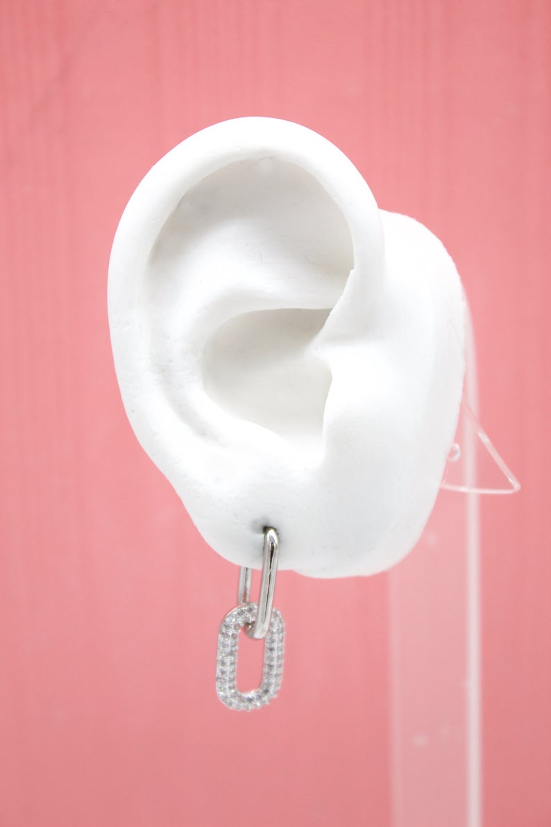 A white mannequin ear displays a silver link hoop earring encrusted with sparkling cubic zirconia stones against a solid pink backdrop.