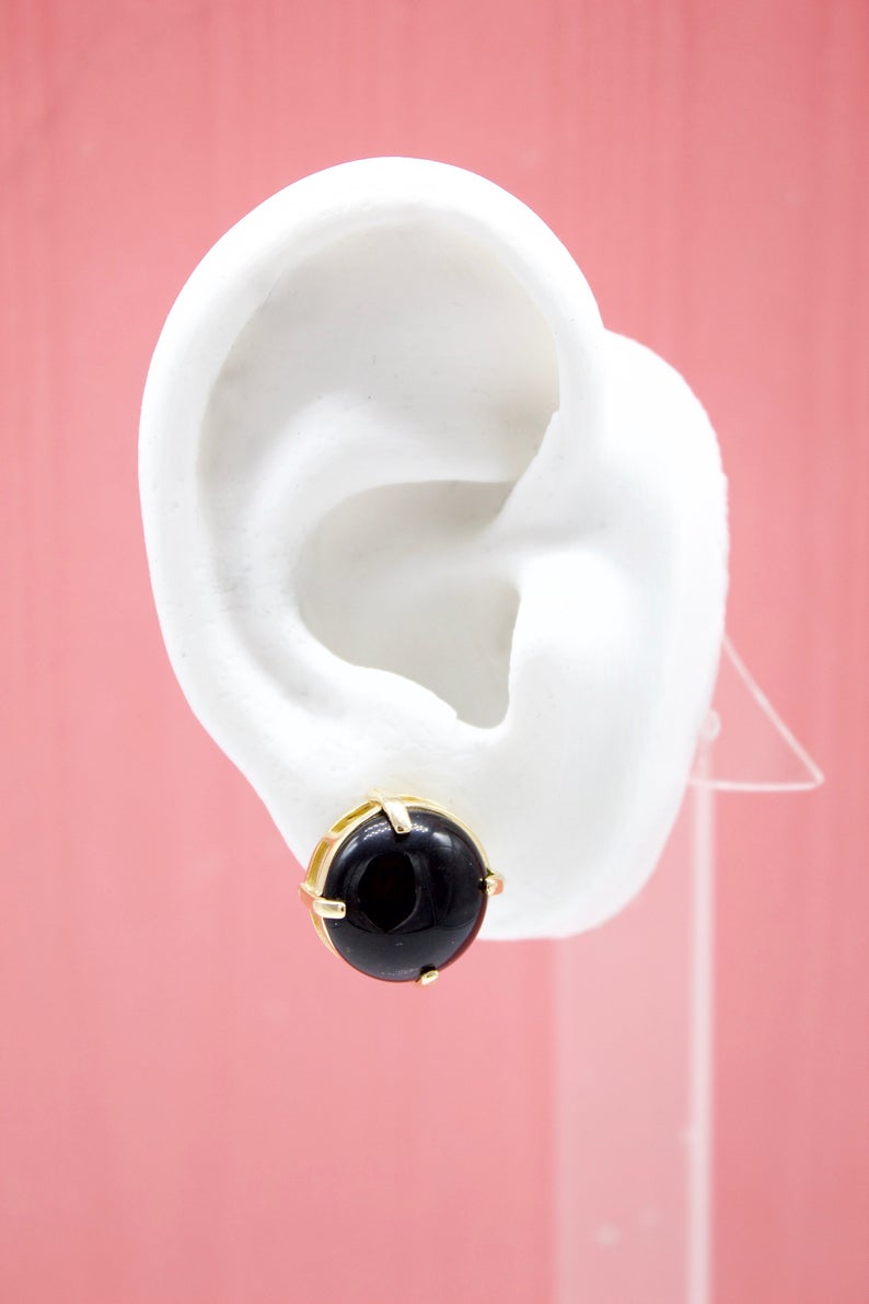 A white mannequin ear displays a round black stud earring set in a gold prongs mounting against a pink background.