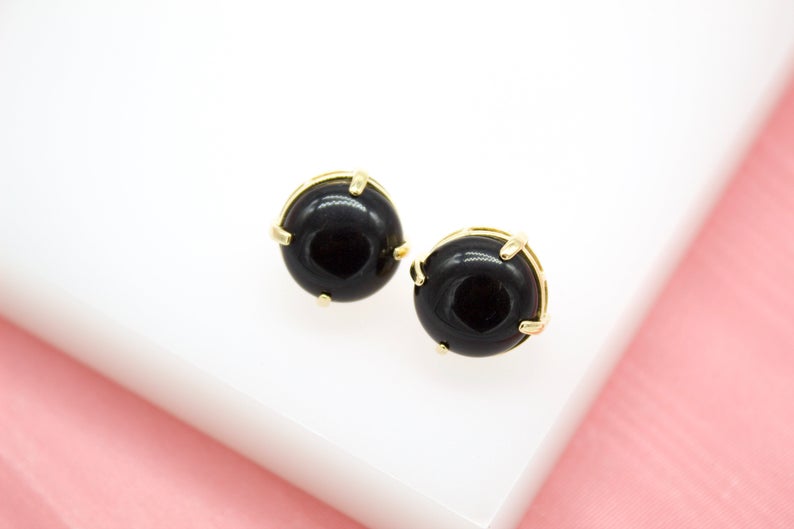 Two round black gemstone stud earrings featuring secure gold prong settings rest on a white display surface.