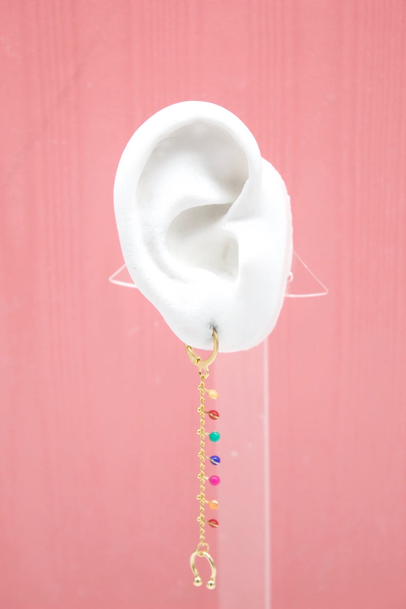 A white mannequin ear displays a gold huggie hoop earring featuring a long, cascading chain adorned with colorful tiny beads against a pink background.