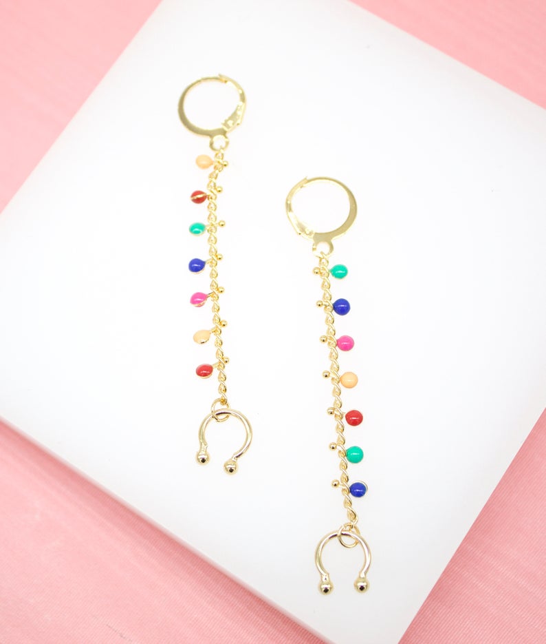 A pair of gold dangle earrings featuring long chains adorned with colorful enamel beads rests on a white display surface.