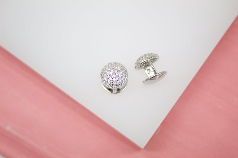 A pair of sparkling silver-toned cuff links with crystal-encrusted domes rest on a clean white display surface against a soft pink background.